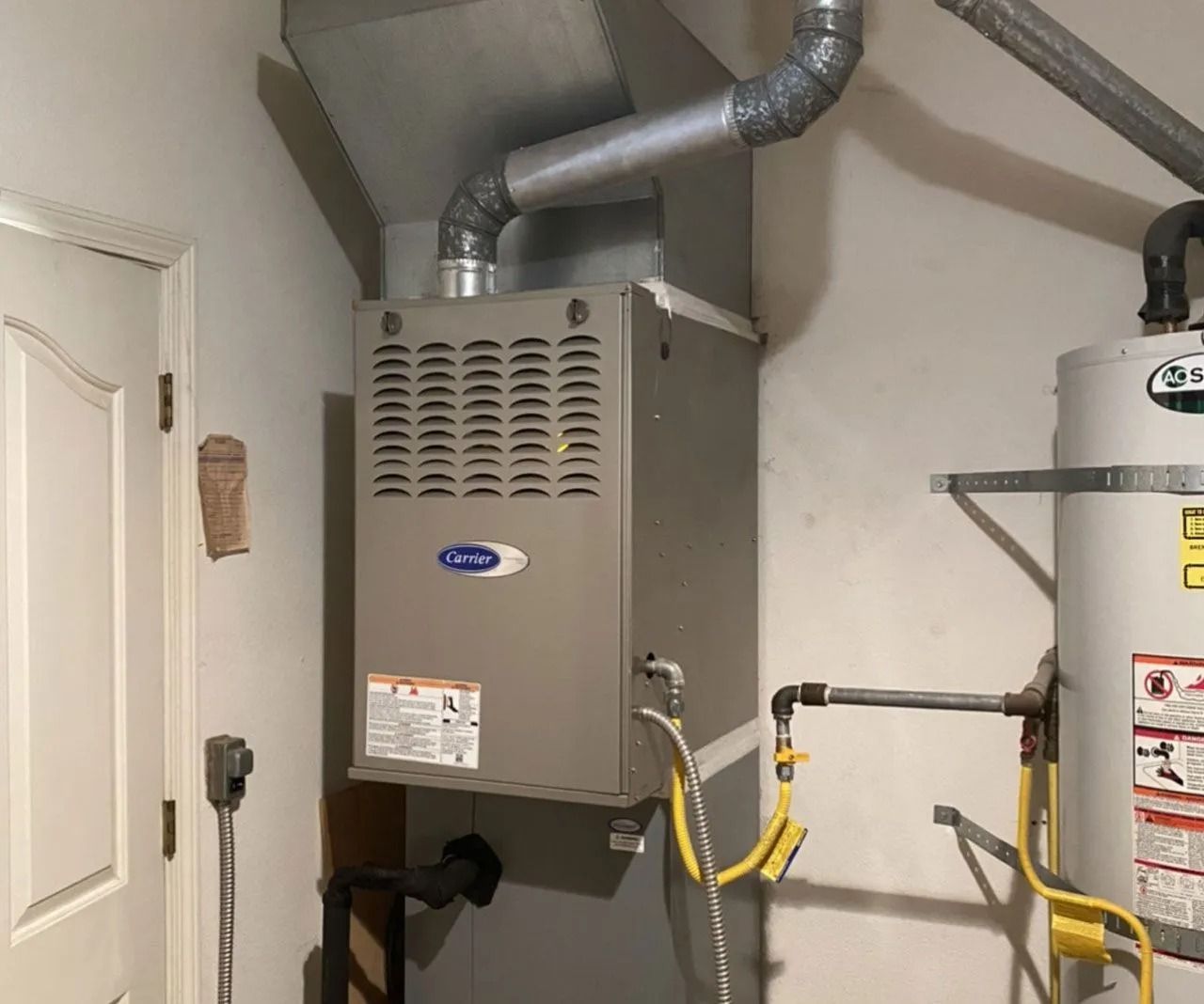A gray furnace mounted on a wall in a utility closet, with ductwork above and a water heater to the right.