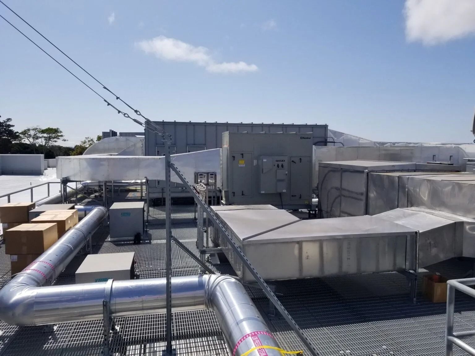 Industrial HVAC rooftop units and metal ventilation ductwork on a flat, gravel-covered roof under a clear blue sky.