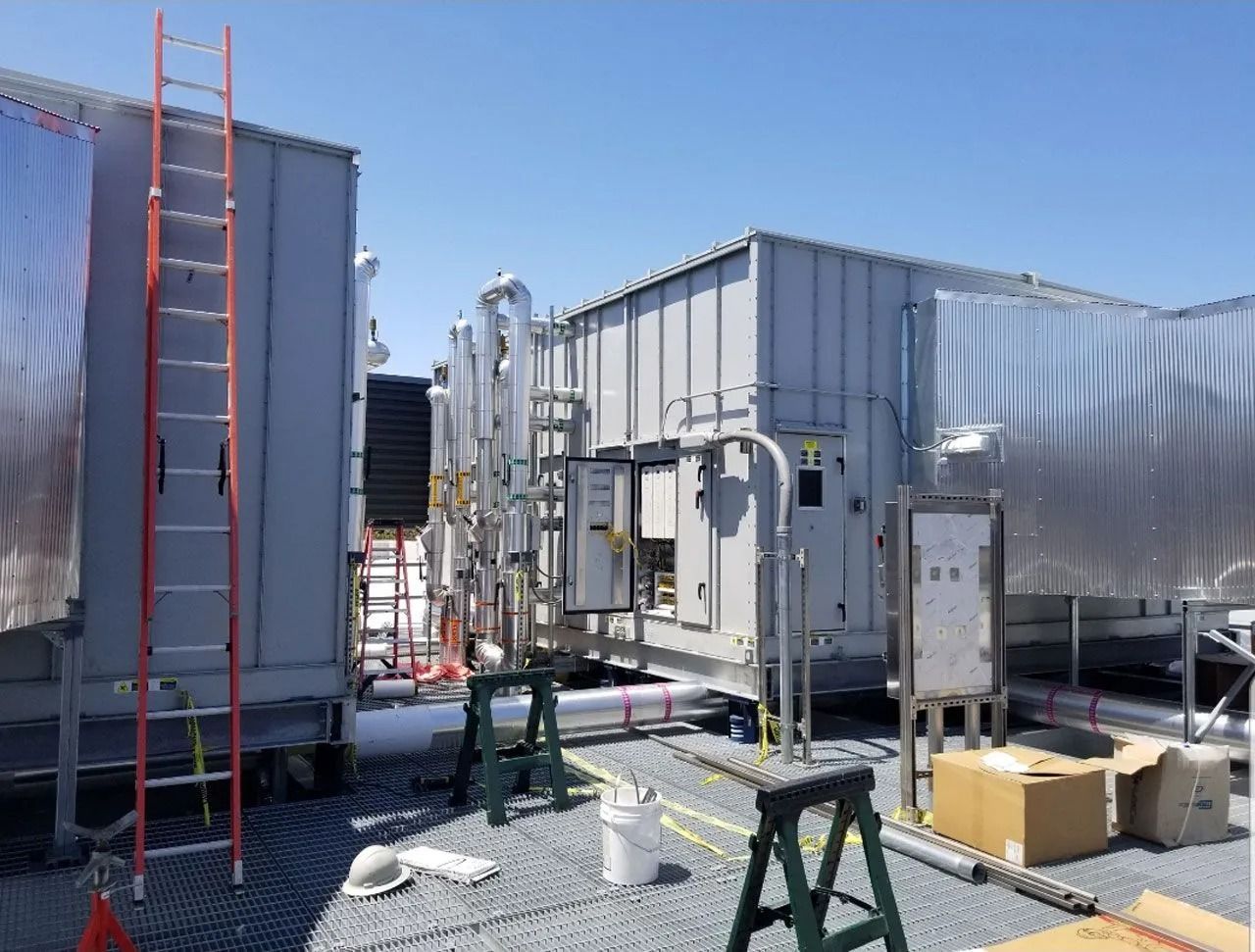 Two large gray HVAC units on a building roof with a ladder, pipes, electrical panels, and construction tools nearby.