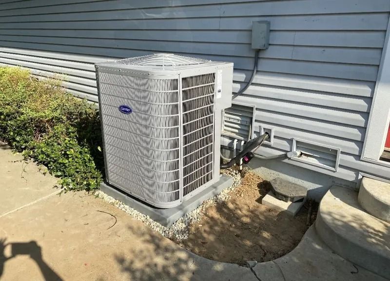 A gray Carrier HVAC unit sits on a concrete pad next to a house with light gray horizontal siding and a bush.