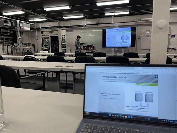 A laptop displaying a piping installation schematic sits in the foreground of a classroom with a presentation on screen.