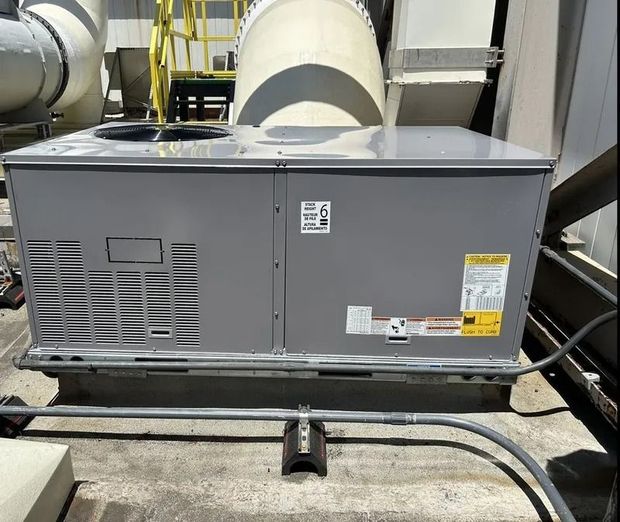A gray, rectangular HVAC rooftop unit sits on a concrete surface, with large ventilation ductwork visible in the background.