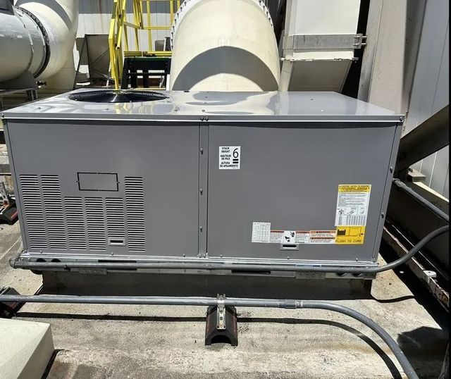 A gray, rectangular HVAC rooftop unit sits on a concrete surface, with large ventilation ductwork visible in the background.