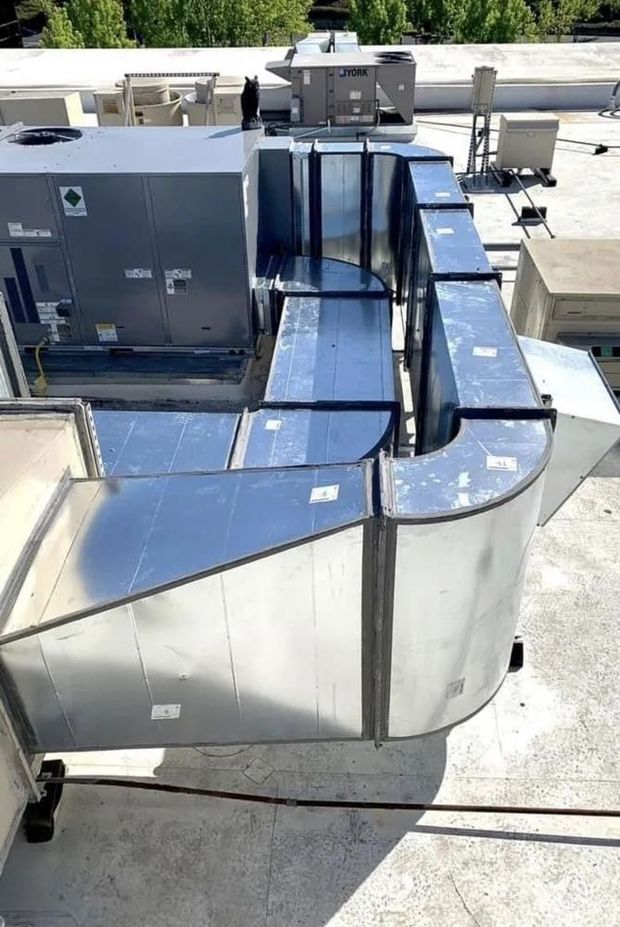 A rooftop view of metal HVAC ductwork extending from a large, grey industrial unit.