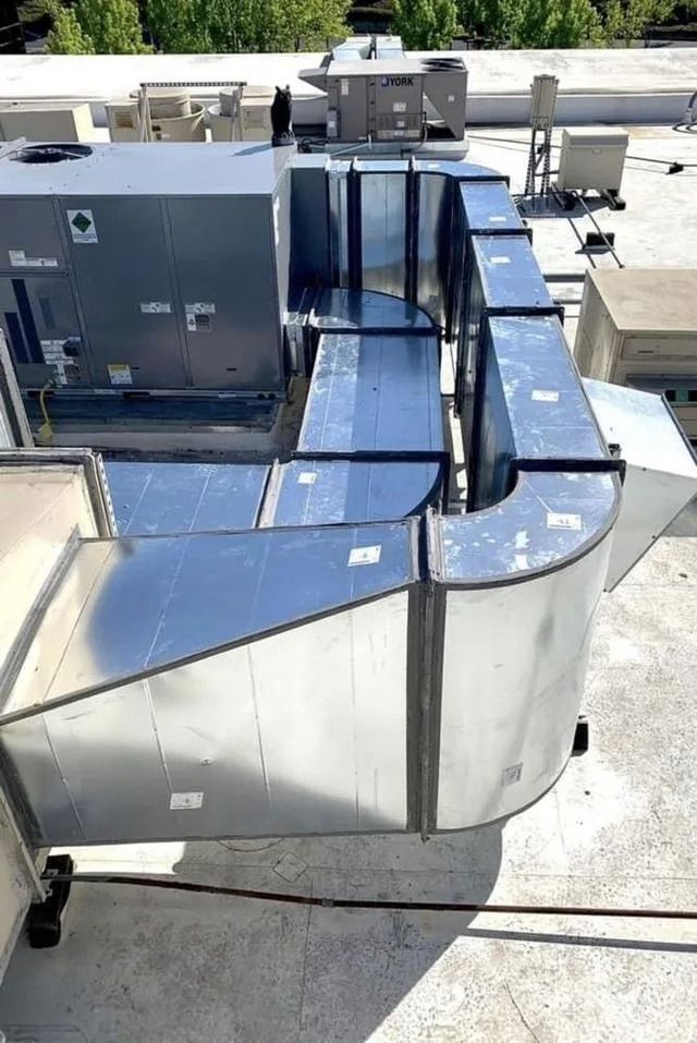 A rooftop view of metal HVAC ductwork extending from a large, grey industrial unit.