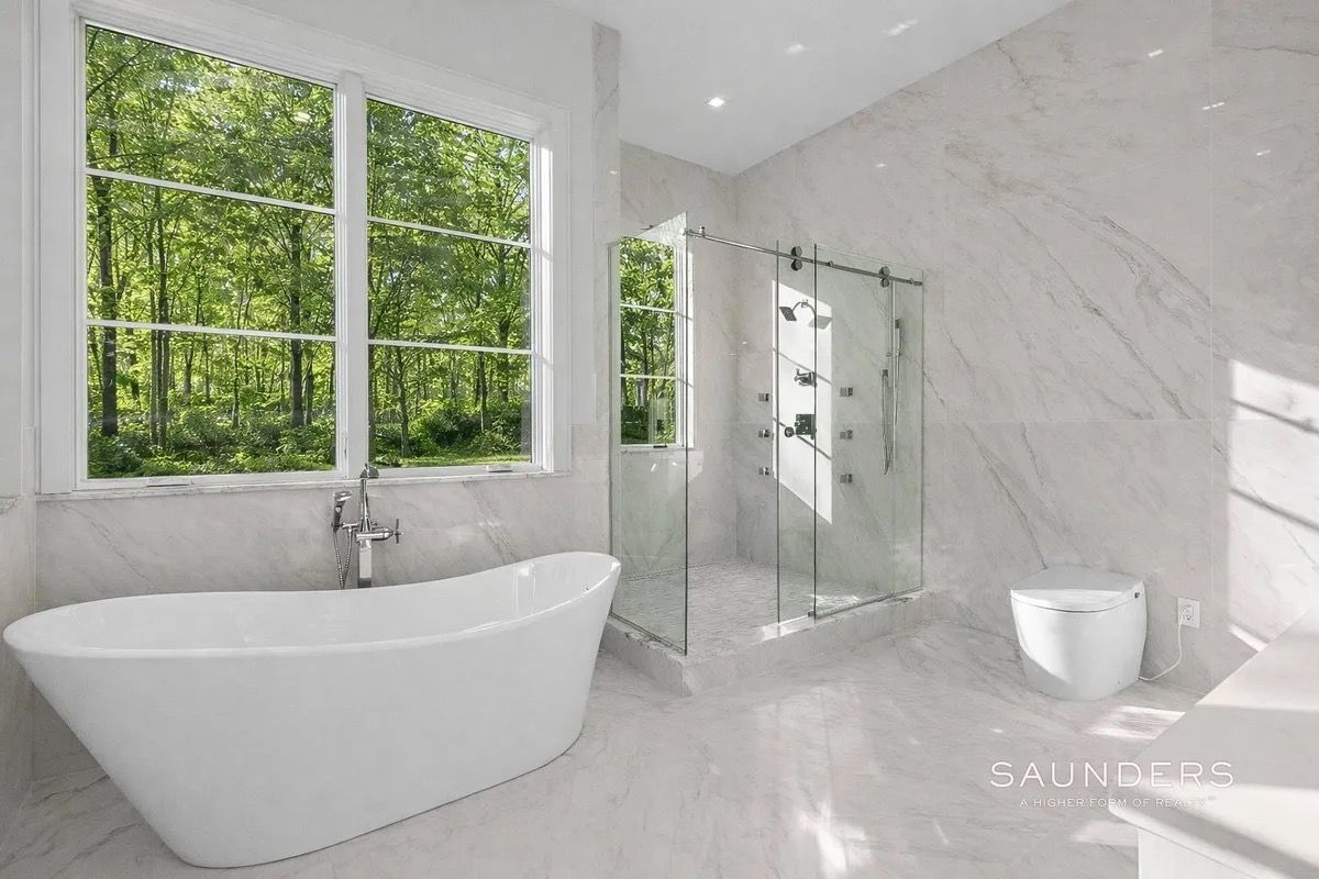Modern white bathroom with freestanding tub, glass shower, marble walls, and large windows overlooking trees