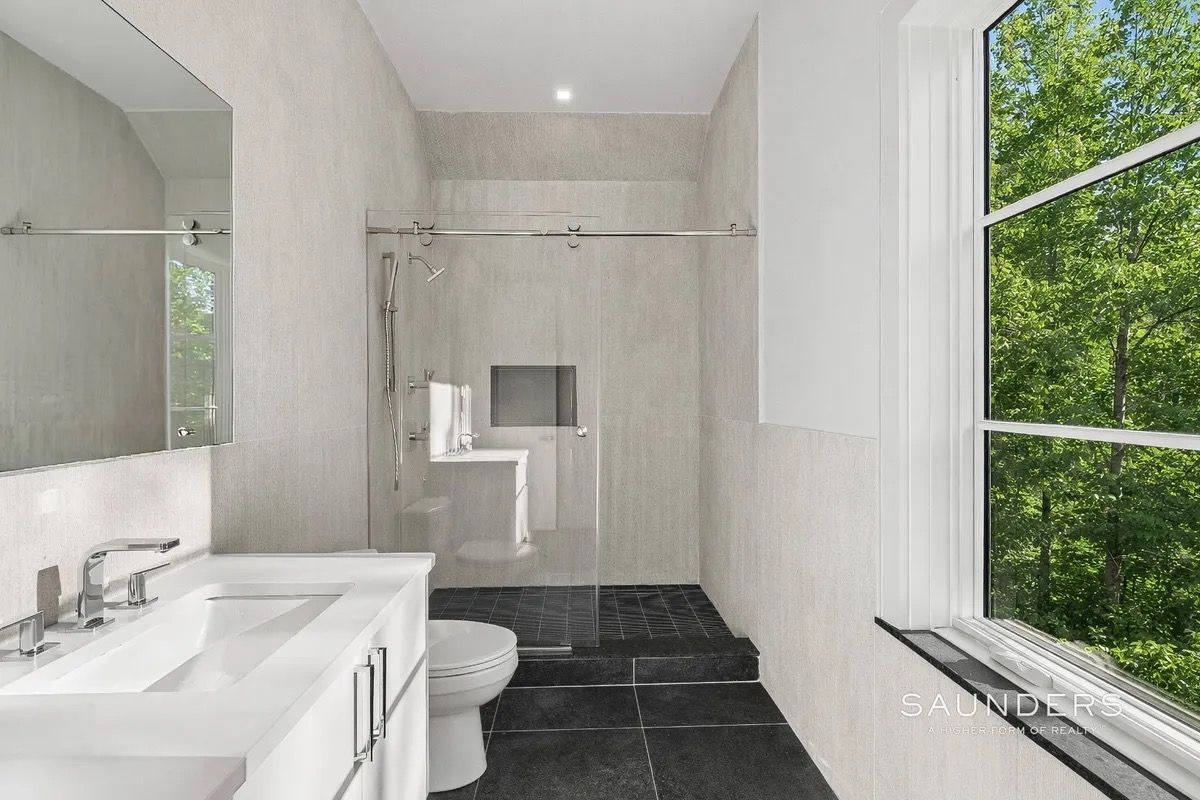 Modern bathroom with white vanity, toilet, walk-in shower, dark tile floor, and large window overlooking greenery