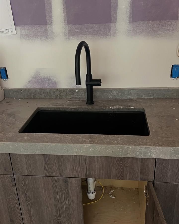 Black kitchen sink with matte black faucet in a gray countertop and wood cabinet base partially open.