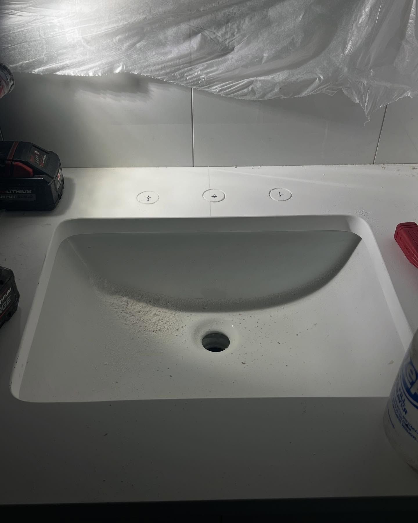 White bathroom sink with central drain and scattered specks, under a plastic-covered mirror or wall.
