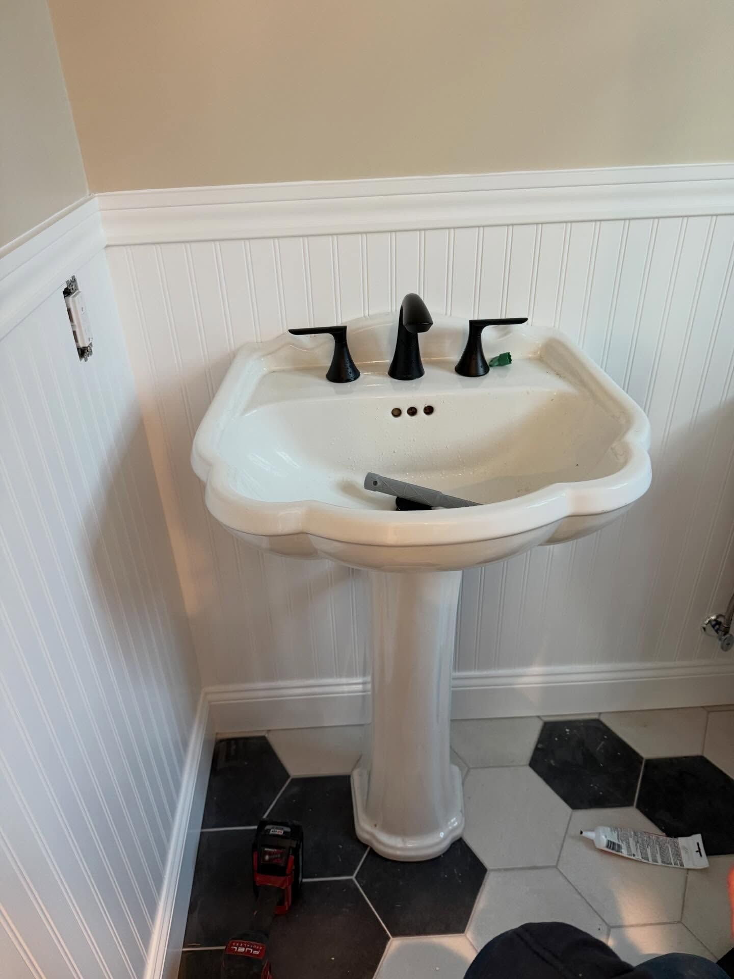 White pedestal sink in a small bathroom with black-and-white hex tile floor and black faucet handles.