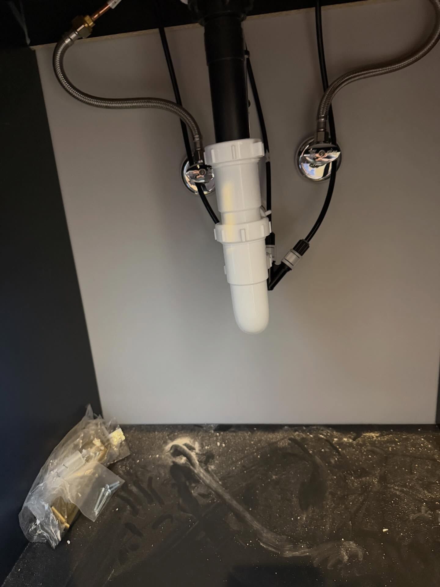 Under-sink plumbing with white drain pipe and black hoses against a gray wall; tools on the floor below.