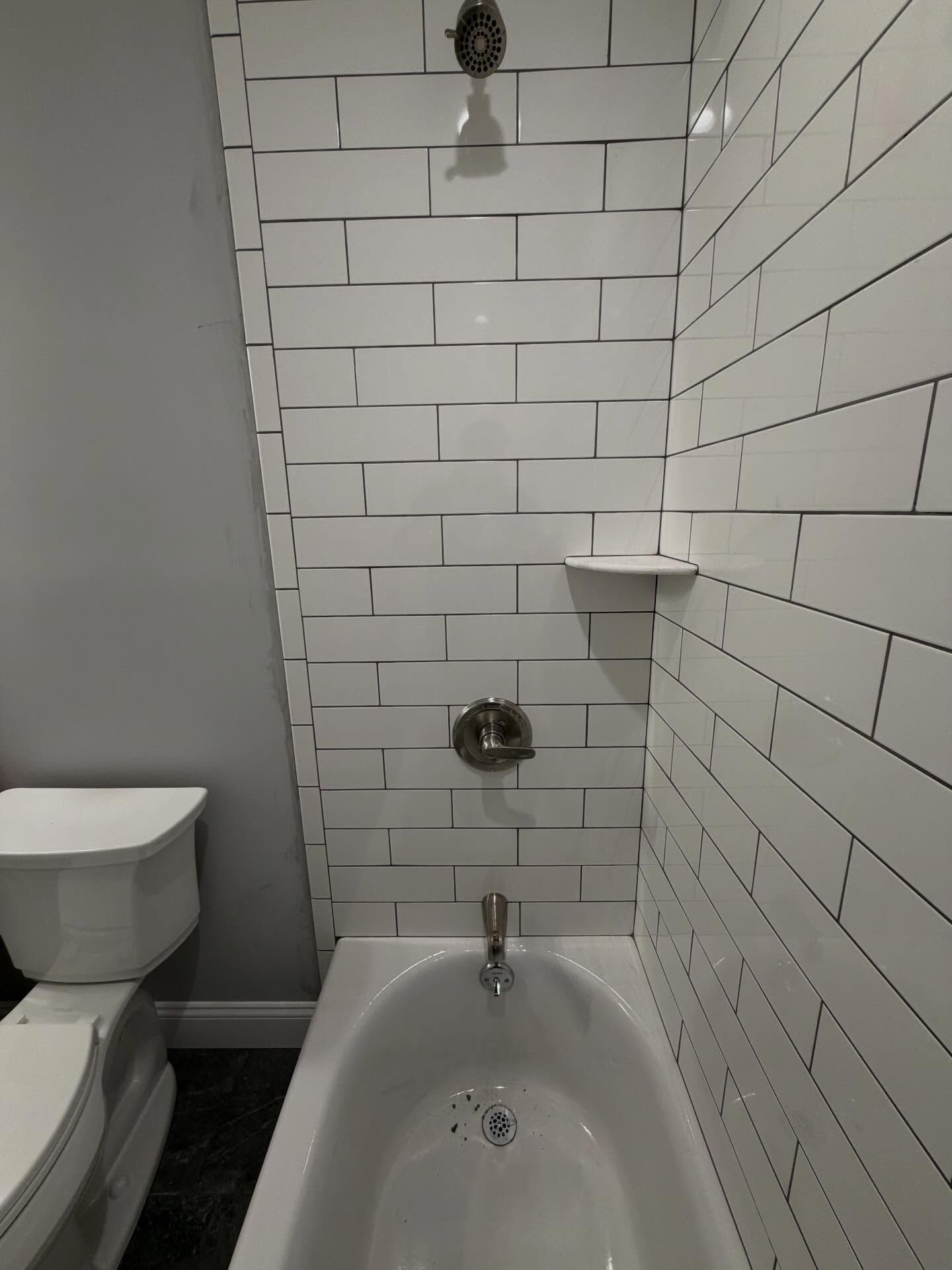 Small bathroom with white subway tile, a bathtub, toilet, showerhead, and wall shelf.