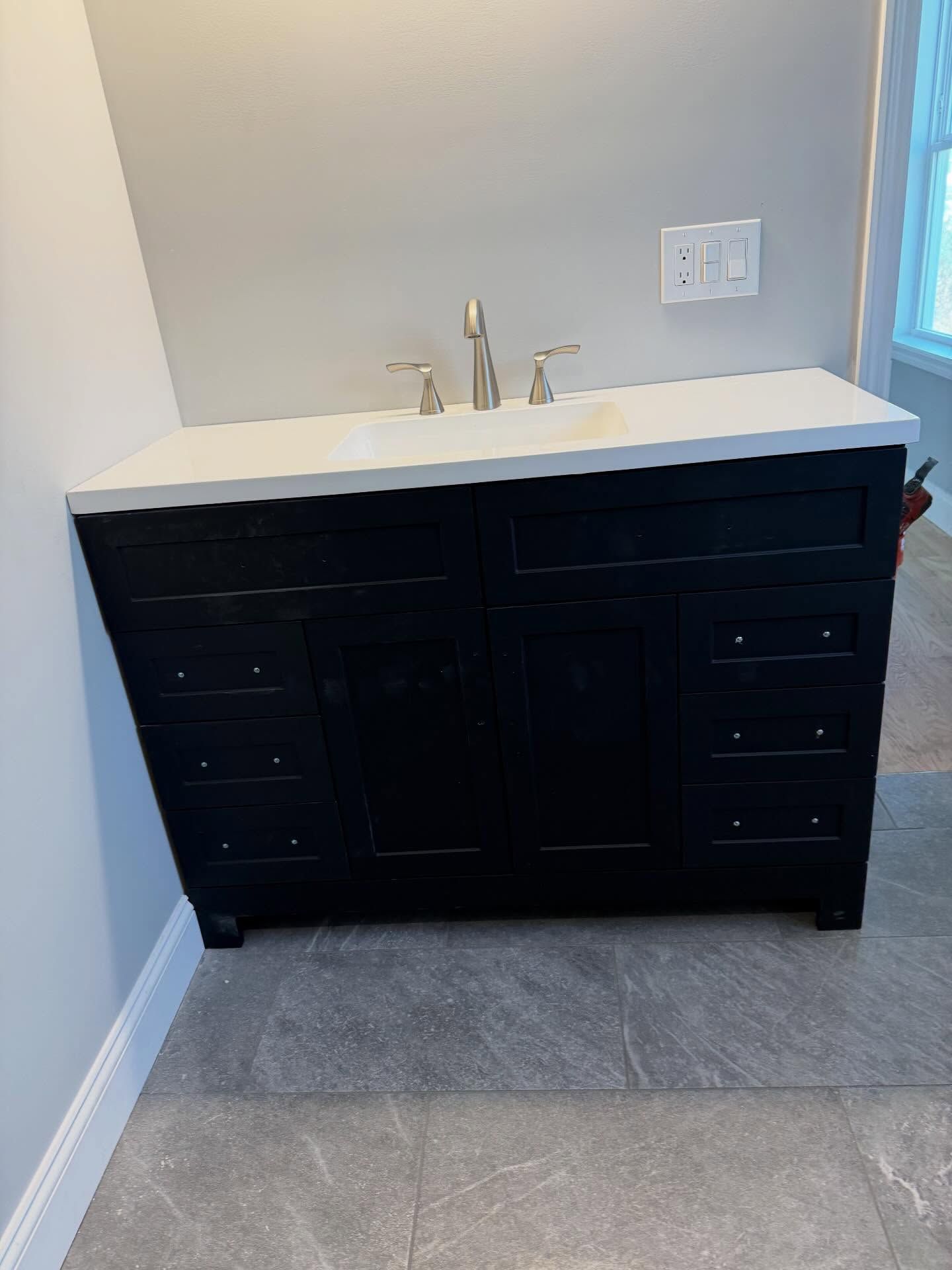 Dark vanity with white countertop and sink in a bathroom corner, gray tile floor, and wall outlets above.