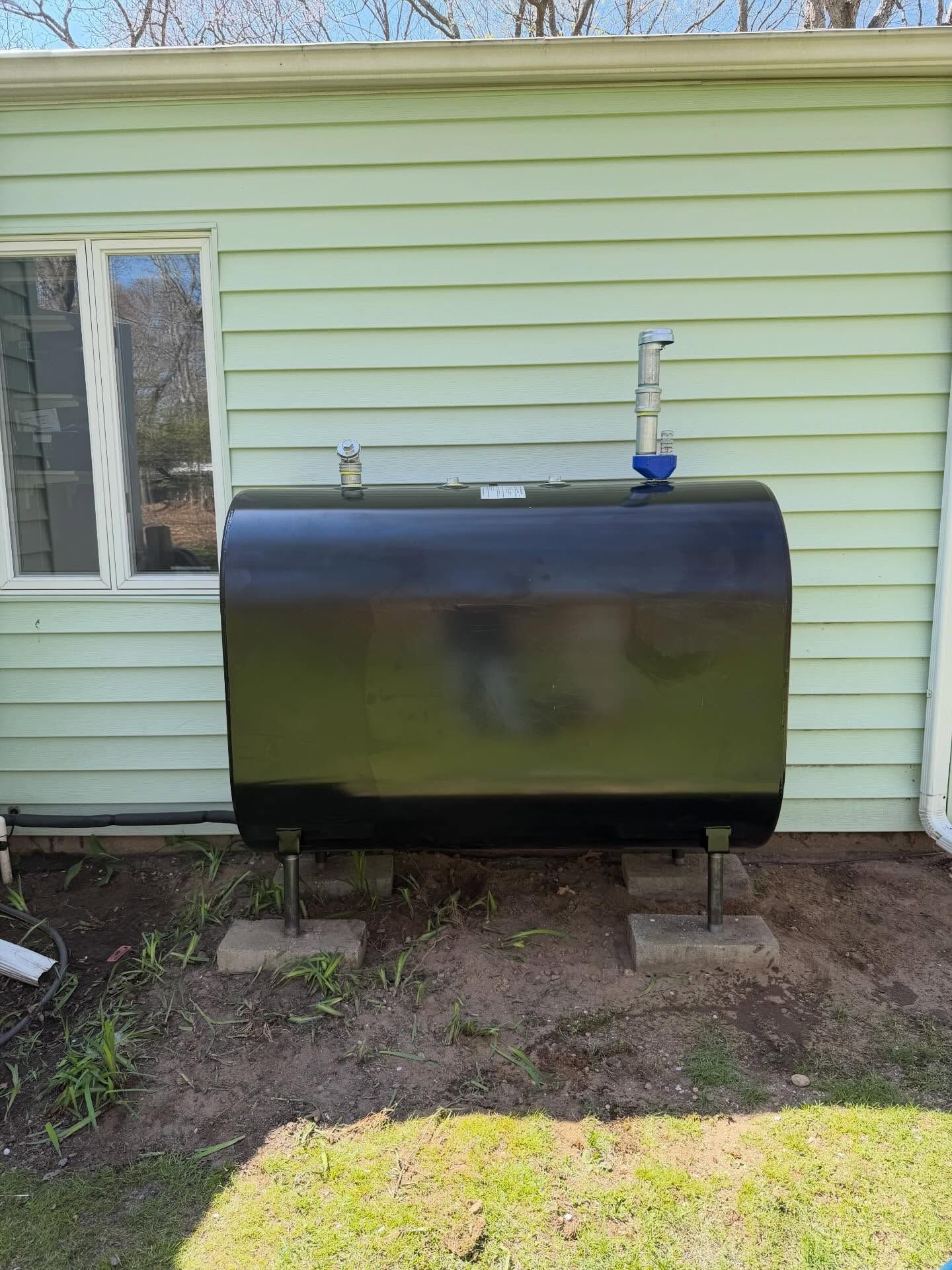 Black tank with pipes against a house exterior, beside a window and dirt yard.