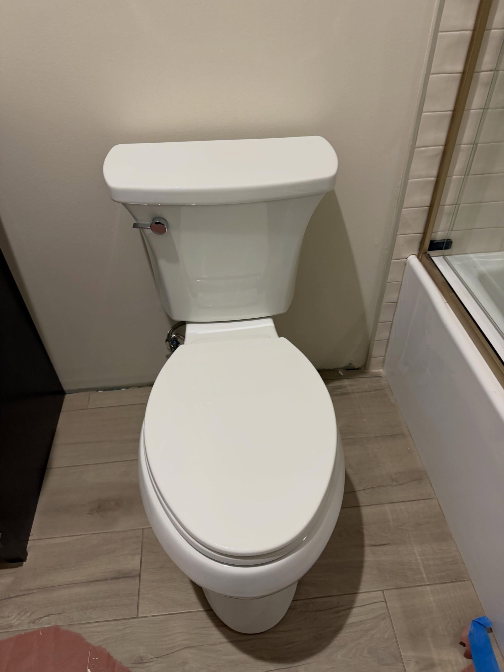 White toilet in a small bathroom beside a shower and light wood-look floor tiles