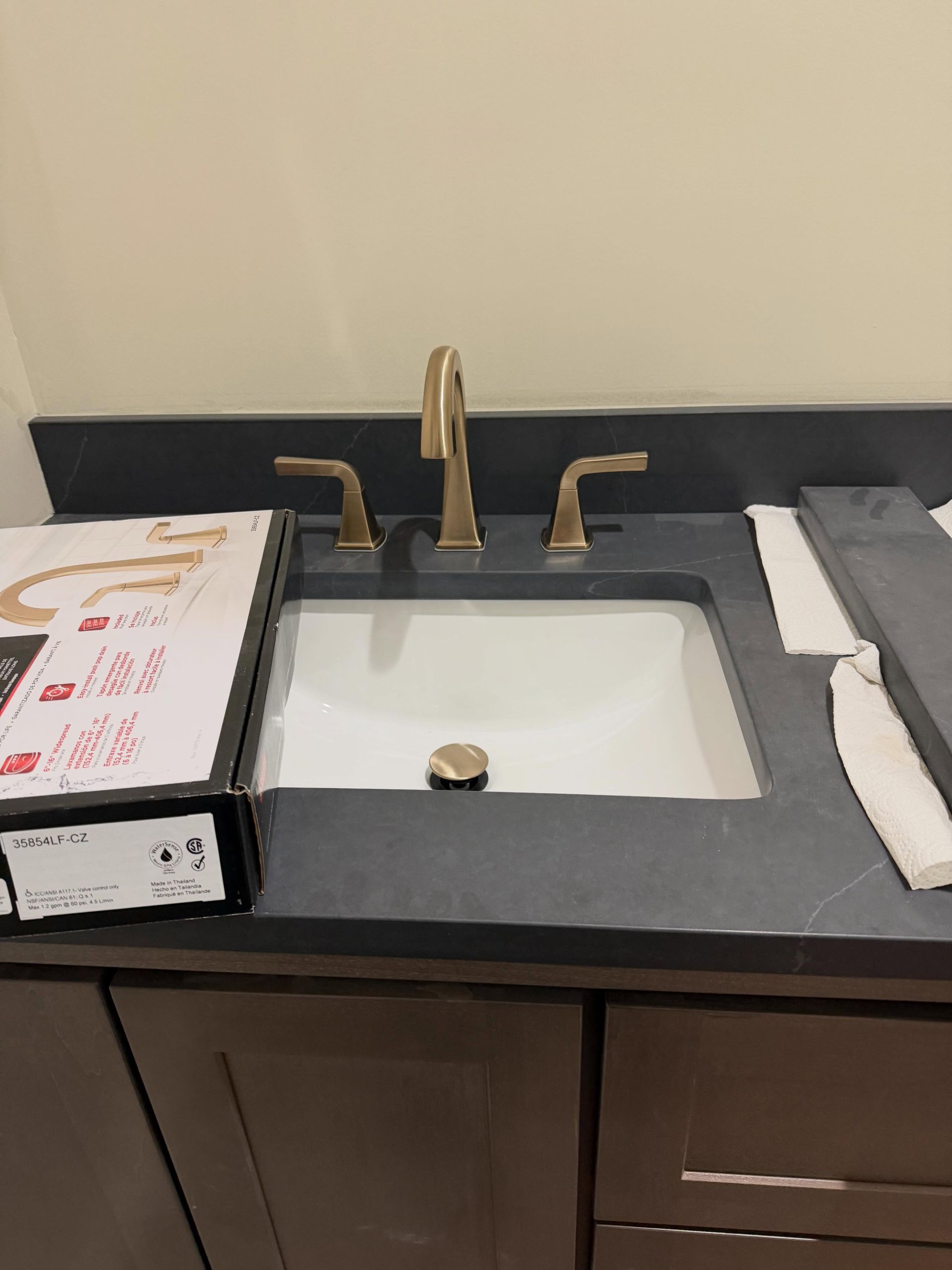 Bathroom sink with dark countertop, gold faucet, and paper towels on both sides.