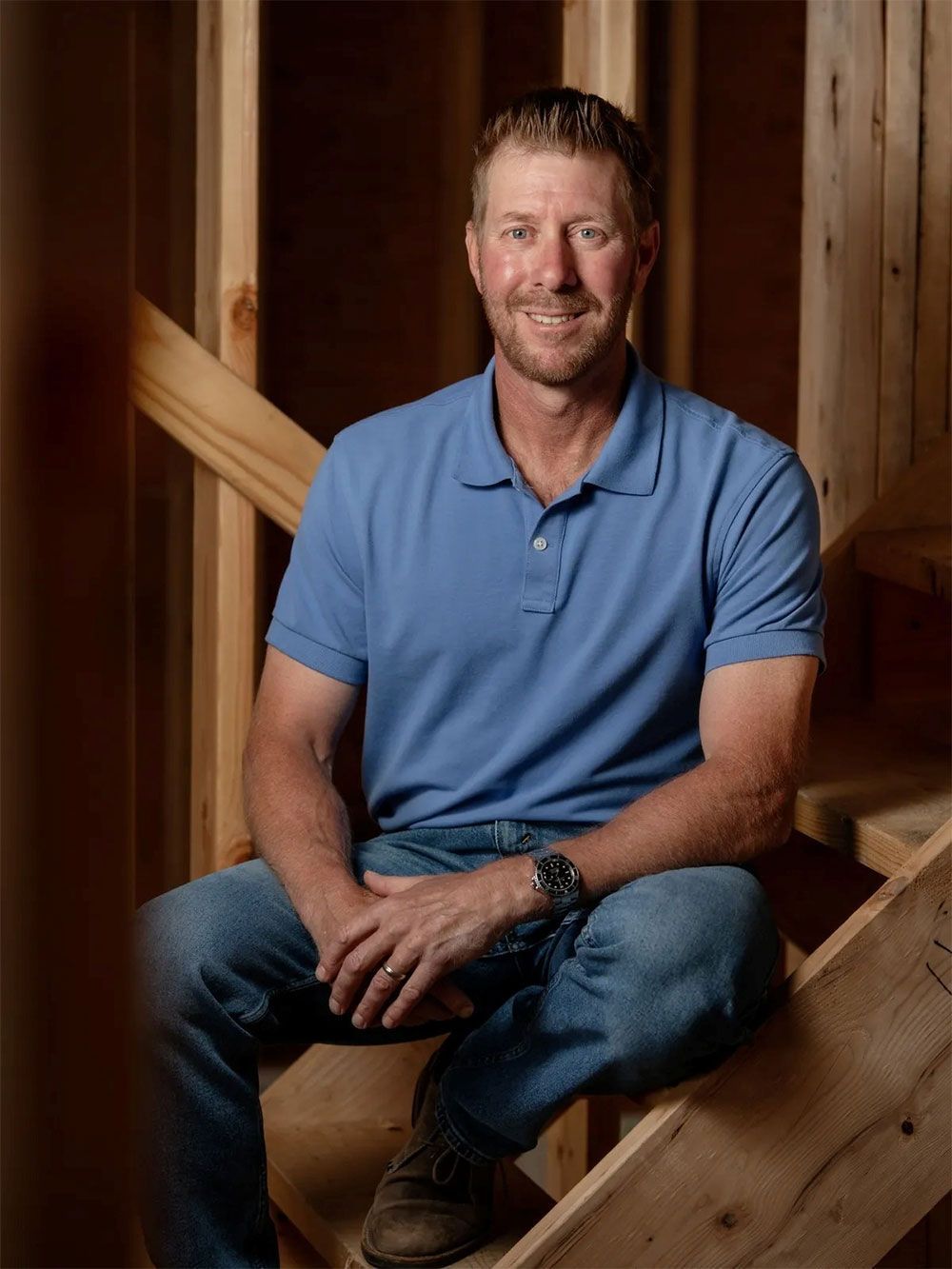 Man in blue shirt and jeans sits on wooden stairs, smiling in a construction setting.