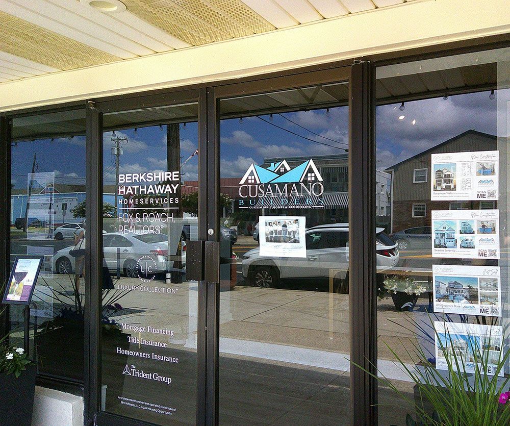 Exterior of a Berkshire Hathaway real estate office with glass doors reflecting the sky, buildings, and cars.