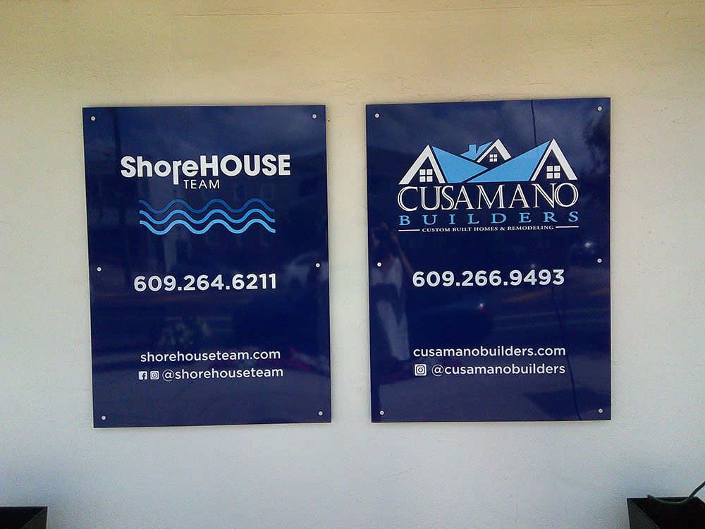 Two blue signs with white text; Shore House Team on left, Cusumano Builders on right, both with contact info and website/social media.