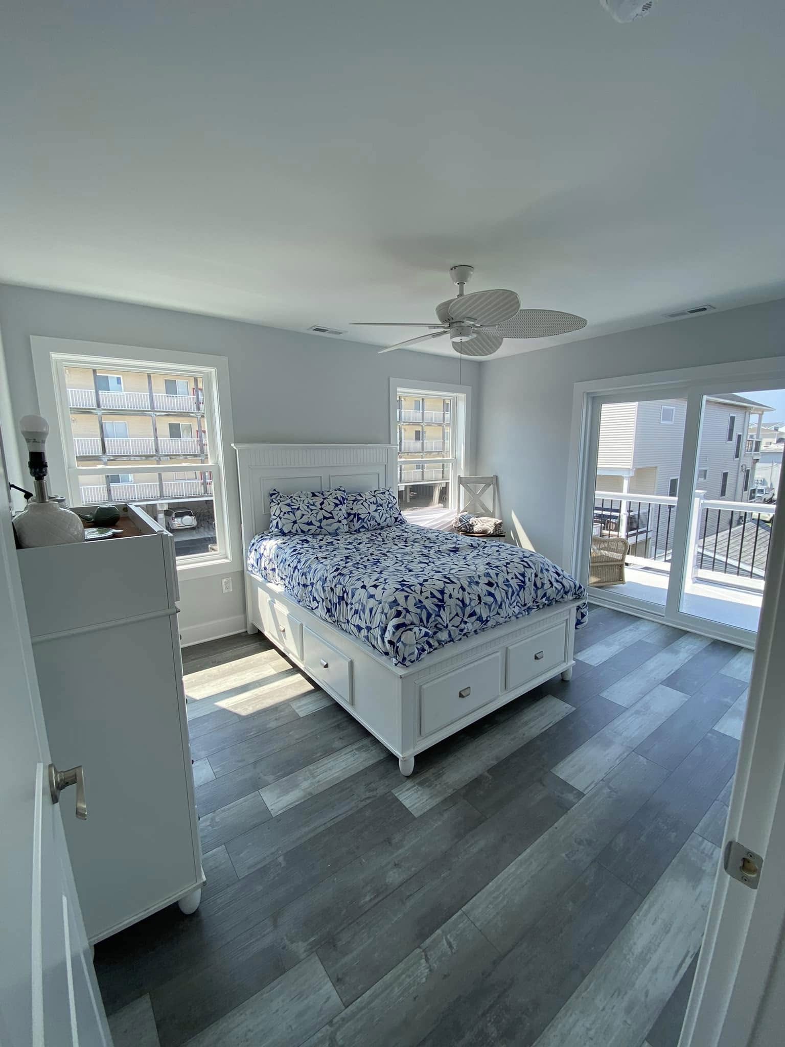 Bedroom with white furniture, blue patterned bedding, gray wood-look flooring, and a view of a sunny deck.