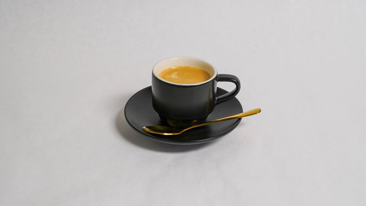 Espresso in a dark gray cup with saucer and gold spoon on a white surface.