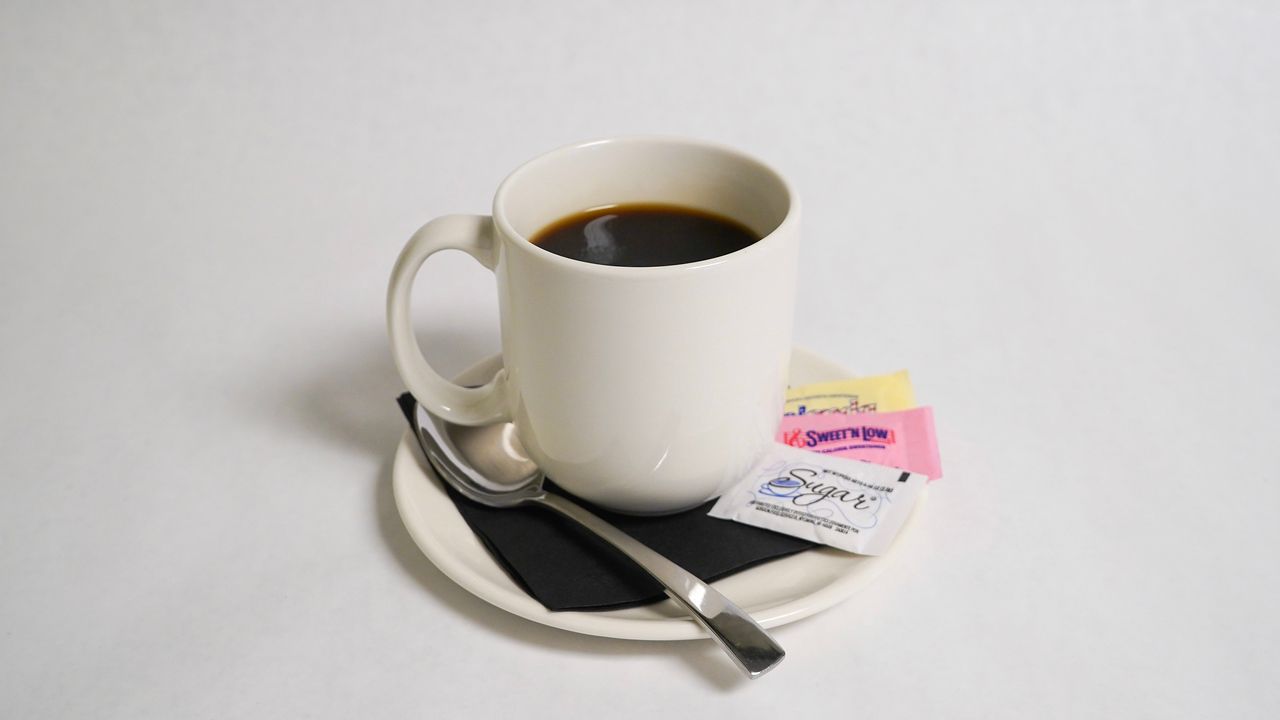 Coffee mug on saucer with spoon, sugar packets, and black napkin on a white surface.