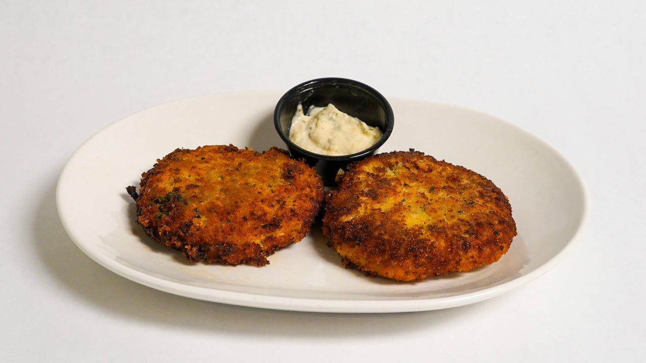 Two golden-brown crab cakes with a small cup of dipping sauce on a white plate.
