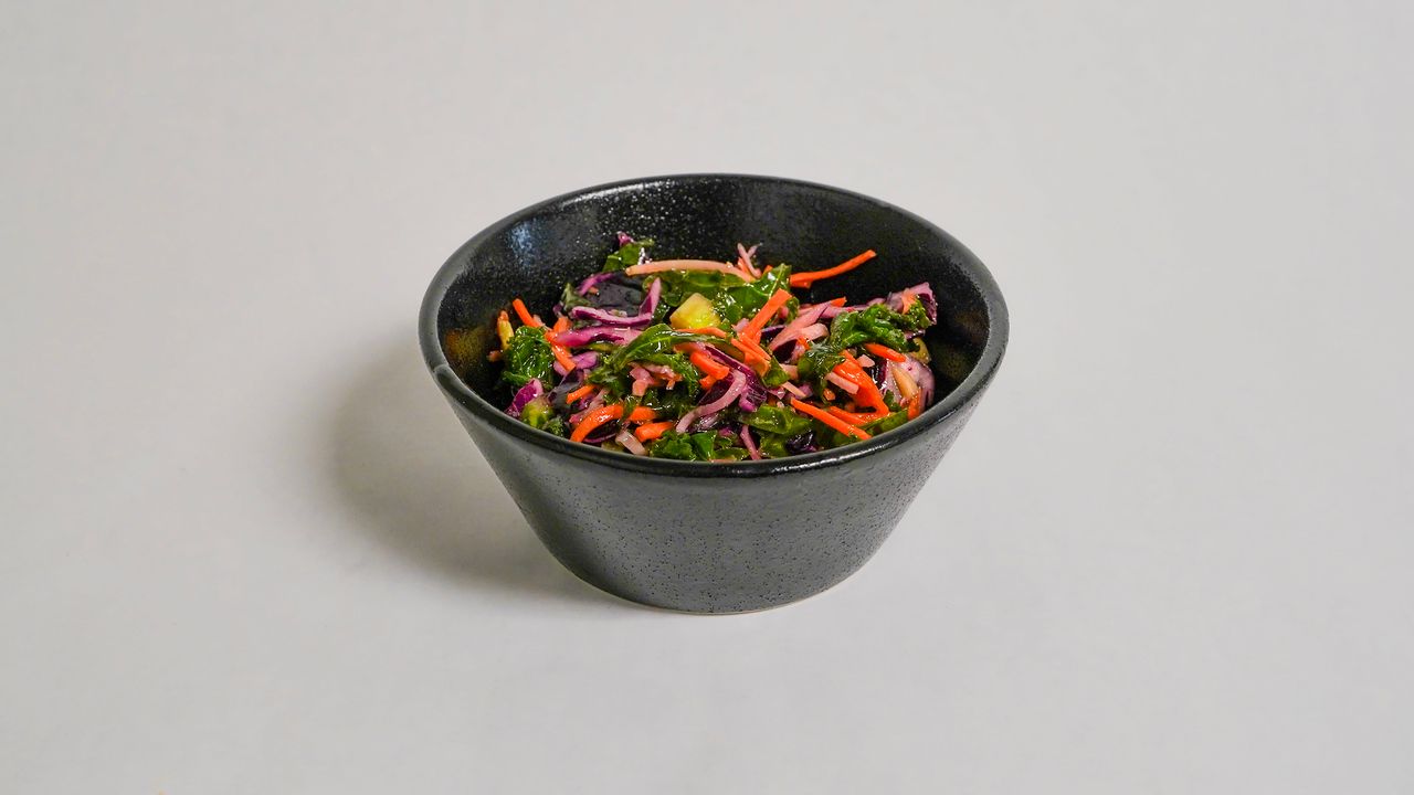 Coleslaw salad in a dark bowl on a white surface. The salad is composed of shredded red cabbage, carrots, and other ingredients.