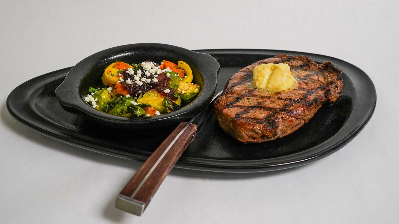Grilled steak with butter, sauteed vegetables, and a knife on a black platter.
