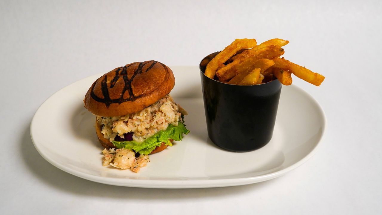 Sandwich with fries on a plate. The sandwich has grilled bun, filling, and lettuce. The fries are in a black cup.