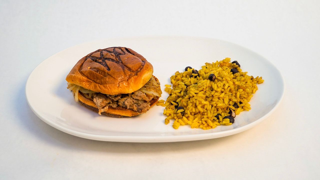 Pulled pork sandwich on a bun and yellow rice with black beans on a white plate.