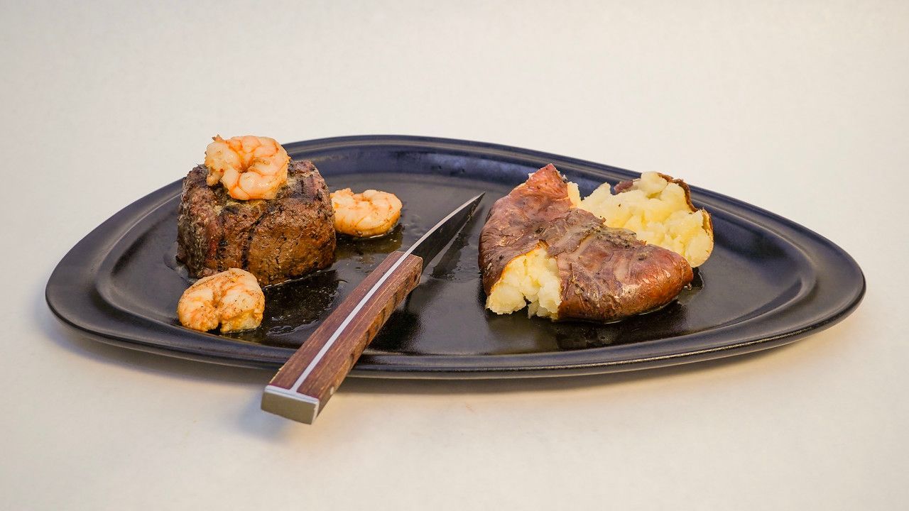 Steak and shrimp with mashed potatoes on a dark blue plate; knife rests across the plate.