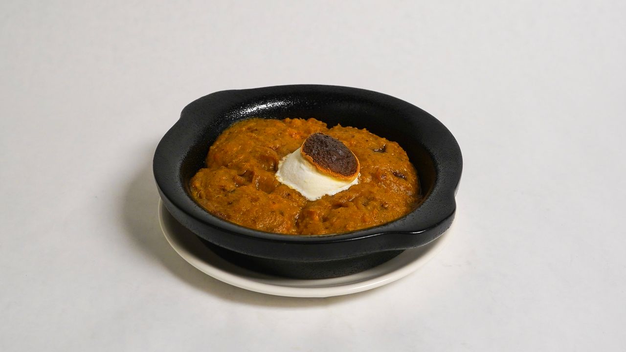 Orange-colored semolina dessert in a black bowl with a scoop of white cream, on a white plate.