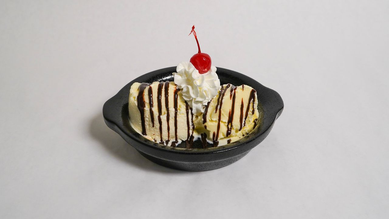 Ice cream sundae in a black bowl, vanilla ice cream, chocolate sauce, whipped cream, and cherry.
