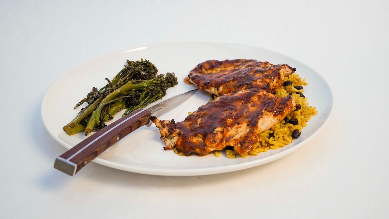 Plate of seasoned chicken with rice, beans, and roasted broccoli. A knife is on the plate.