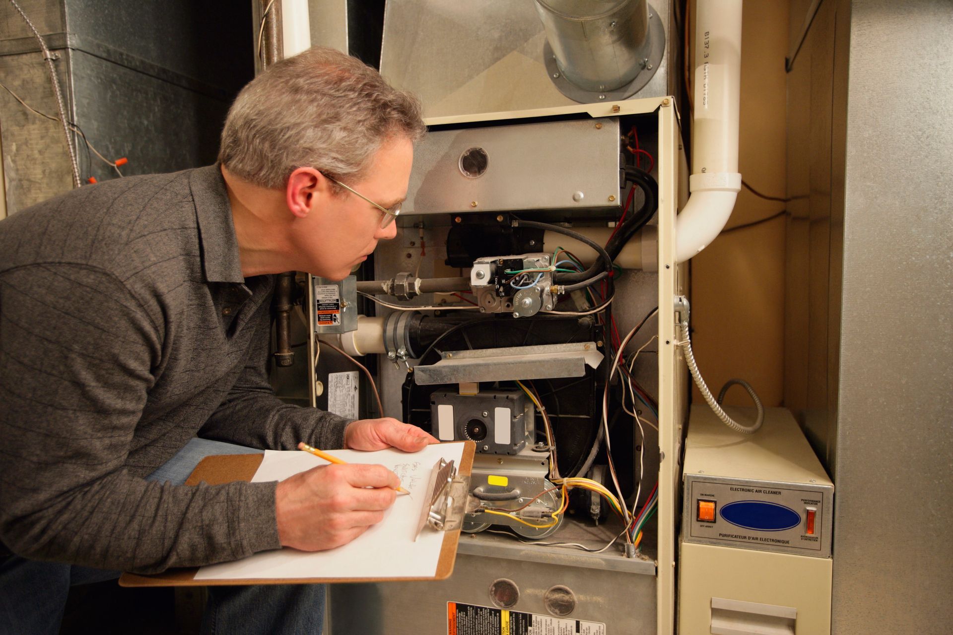 Technician inspecting an open furnace unit with clipboard and tools in a utility room