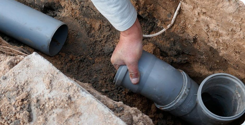 Worker fitting gray PVC pipe in a dirt trench beside an open pipe opening.