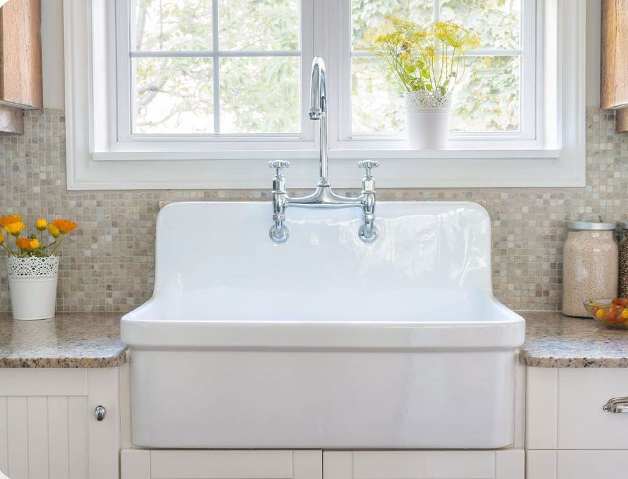 White farmhouse sink beneath a window, flanked by potted flowers and glass jars in a bright kitchen