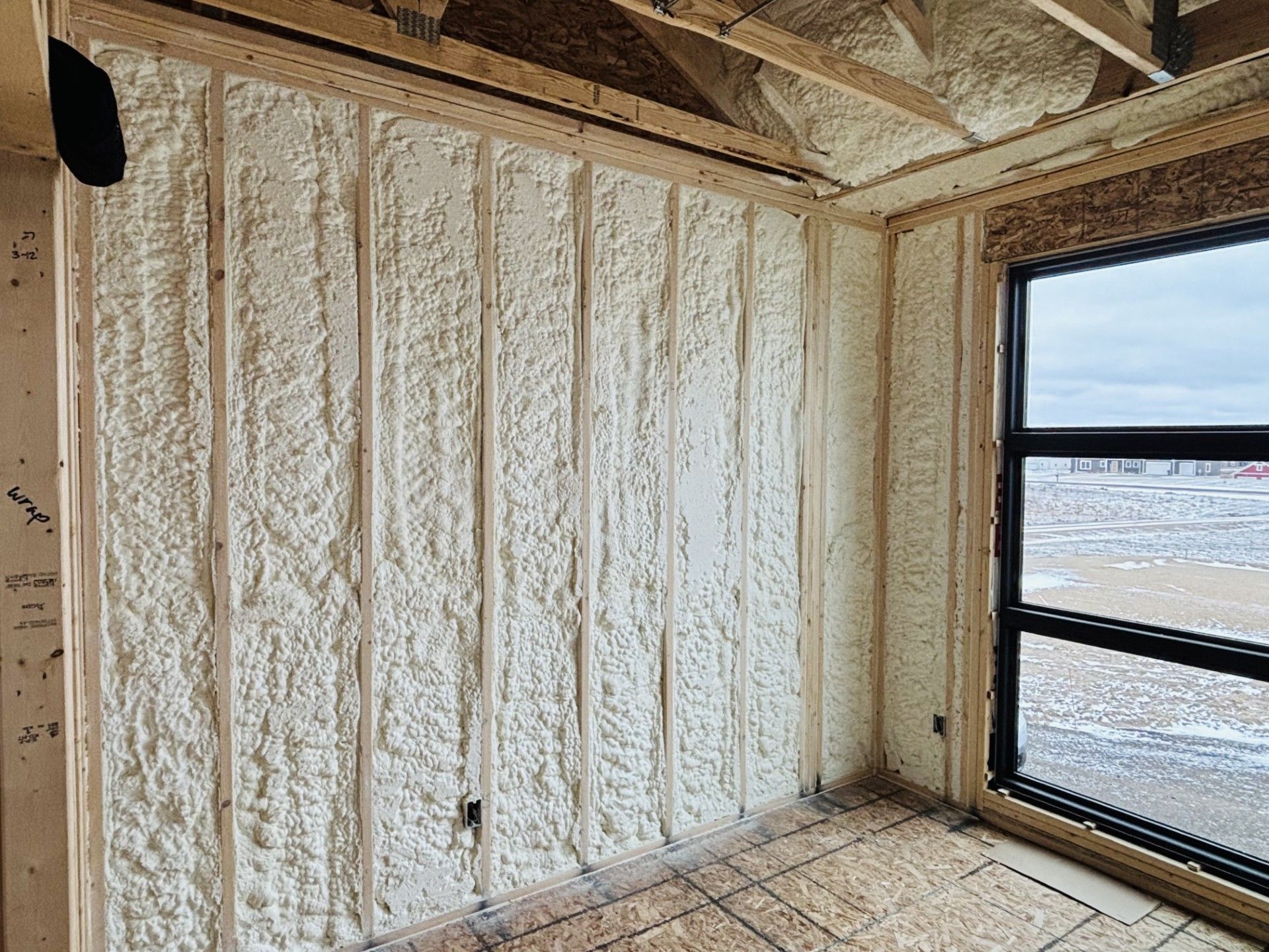 Interior of a room under construction, with spray foam insulation on walls and ceiling, near a window.