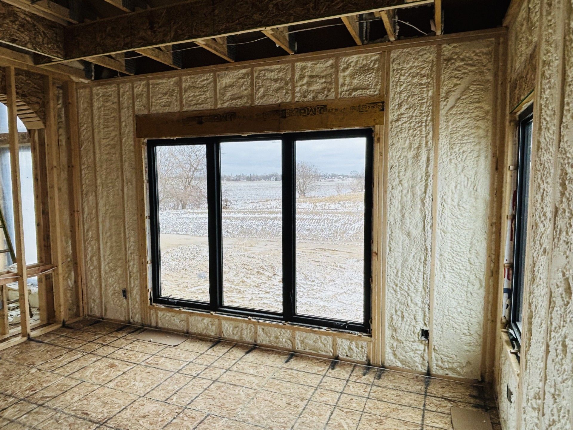 Interior view of a room under construction with exposed wooden framing and spray foam insulation. A large window overlooks a snowy outdoor scene.