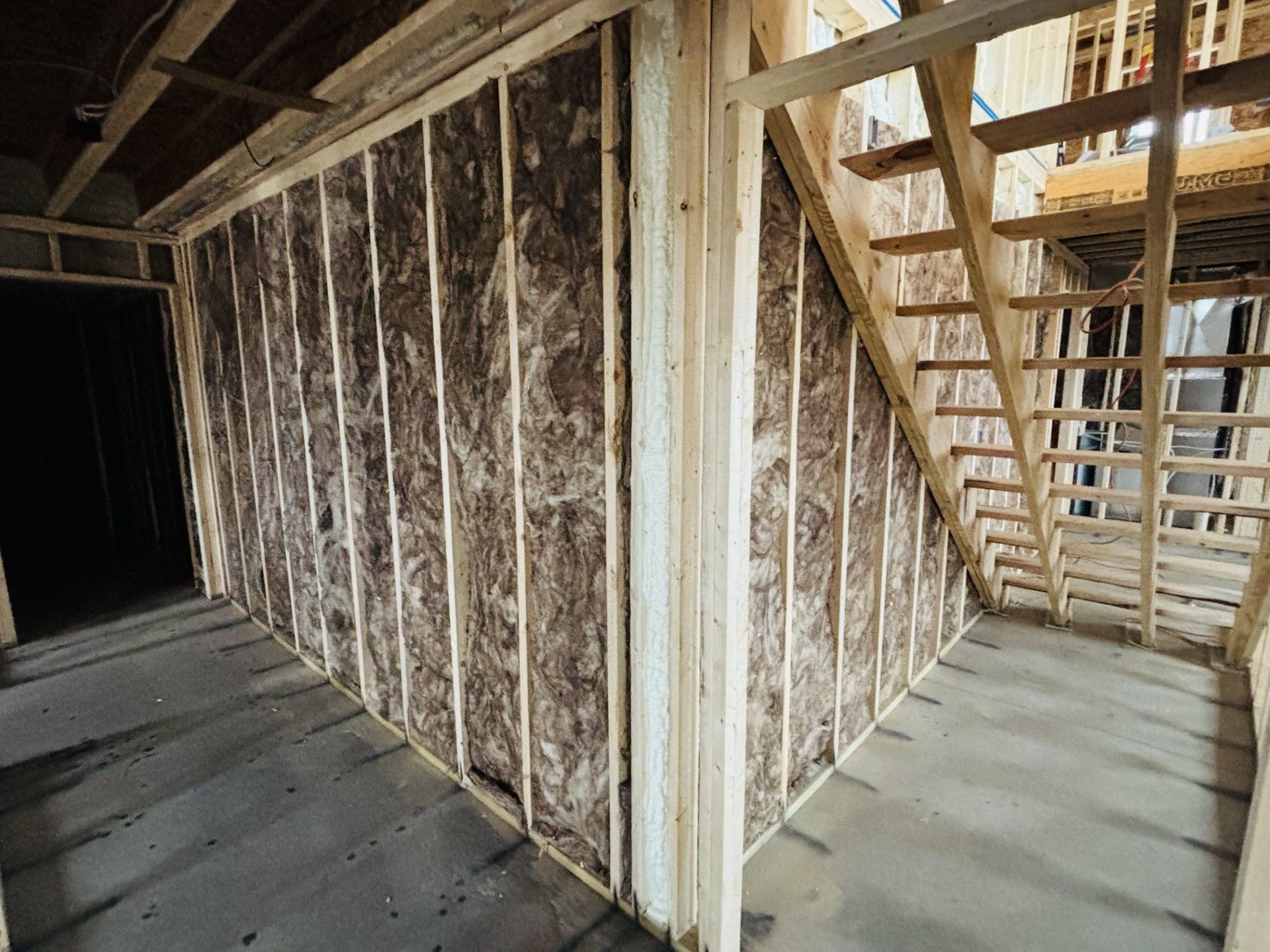 Interior construction with insulated walls and staircase framing. Grey concrete floor, wooden studs, brown insulation.