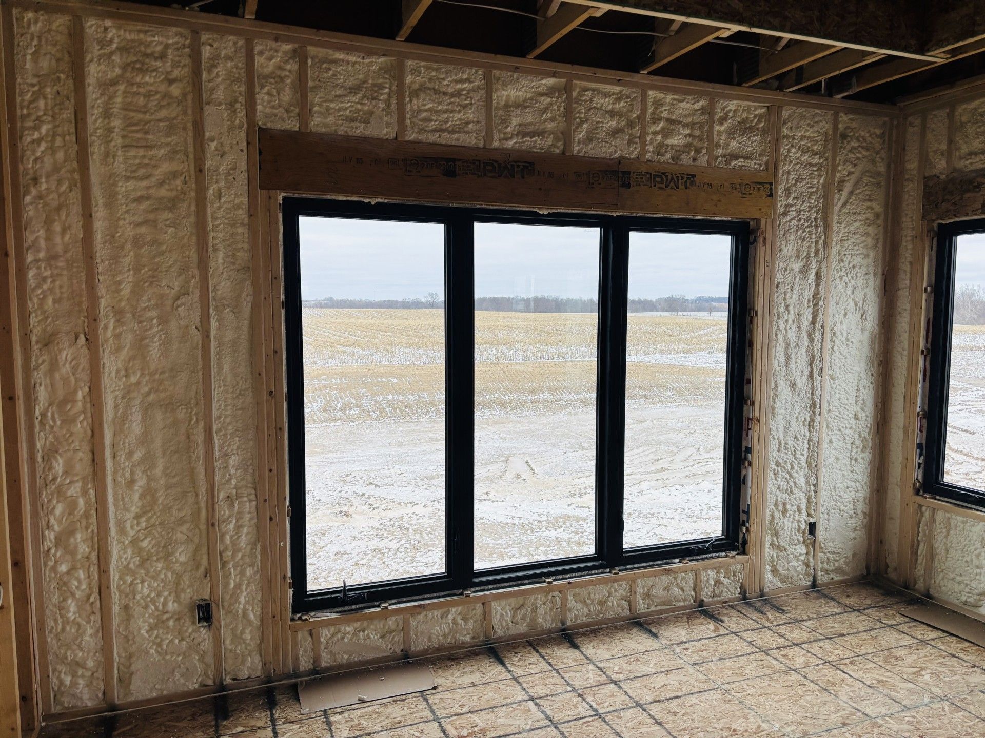 Interior view of a room under construction with spray foam insulation on the walls and a window overlooking a snowy field.