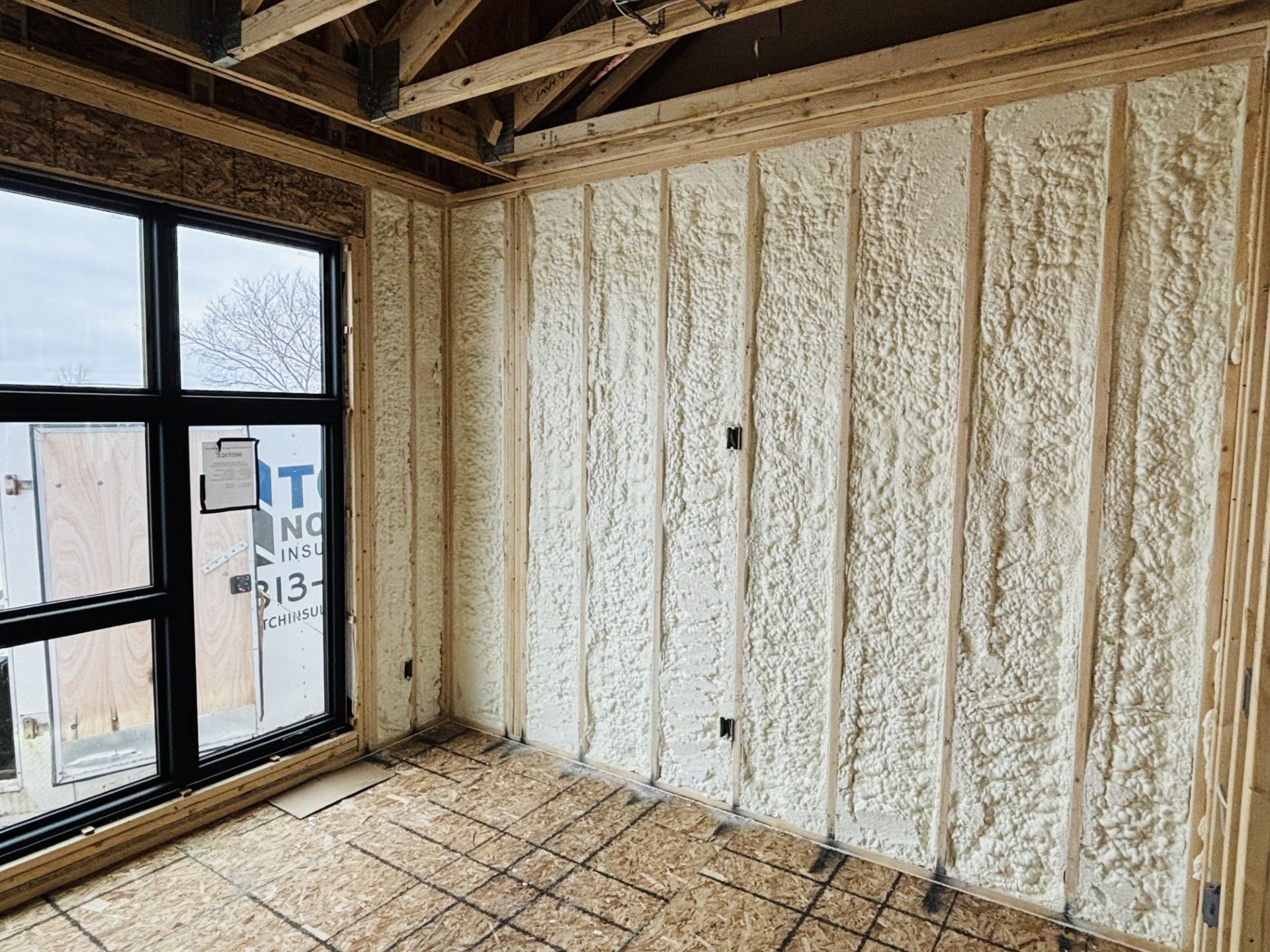 Interior room under construction, spray foam insulation on walls, window on left, wooden framing.