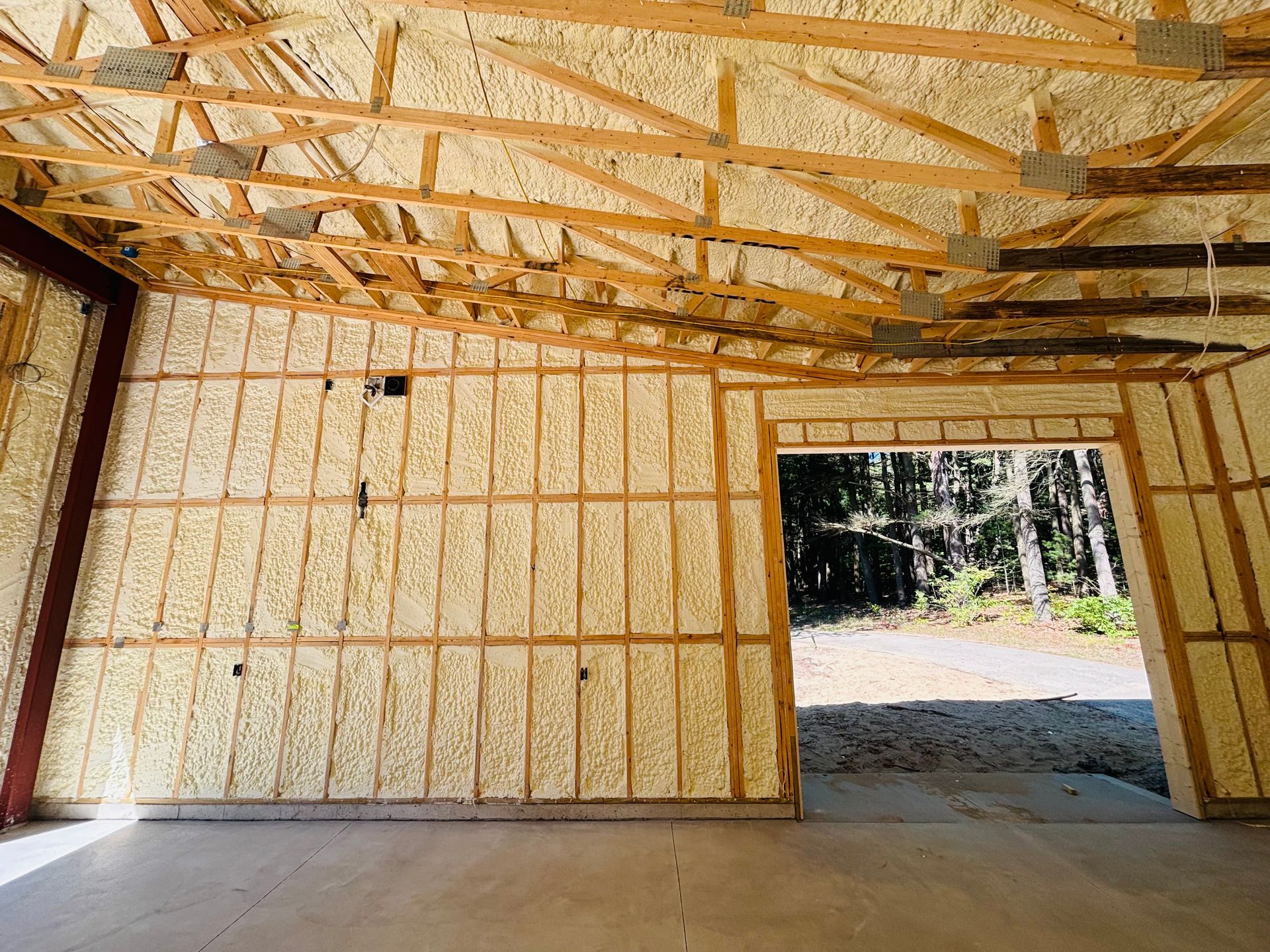 Interior of a building under construction, walls and ceiling insulated with yellow spray foam, open doorway with view of trees.