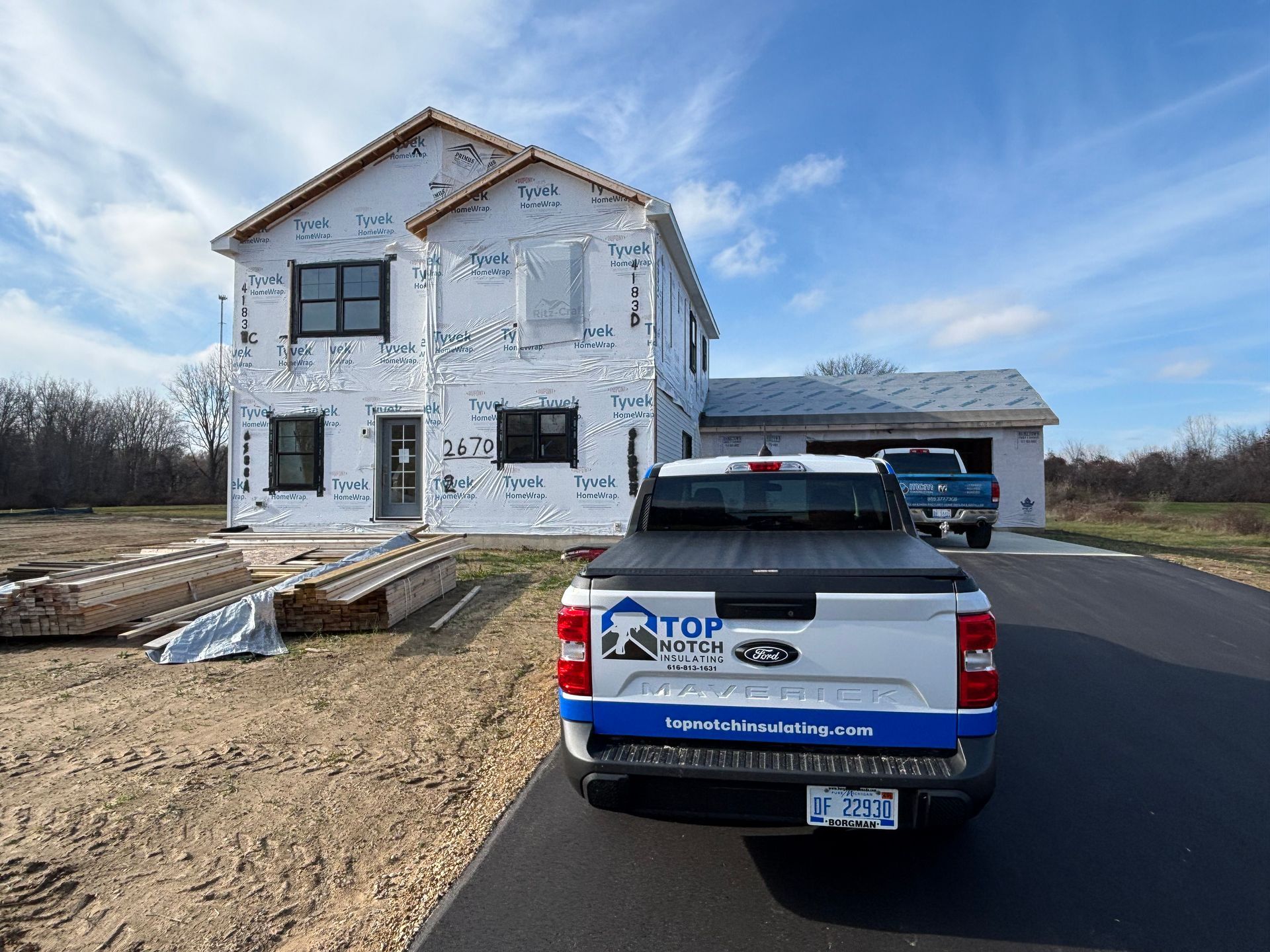 Construction site with two-story house wrapped in Tyvek, pickup truck parked on asphalt driveway.