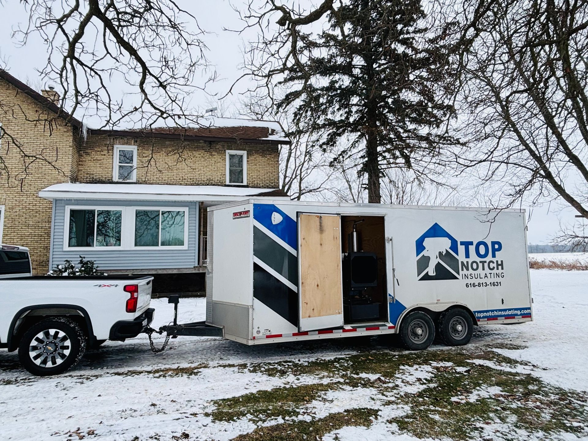 Truck towing a trailer parked near a house covered in snow. 