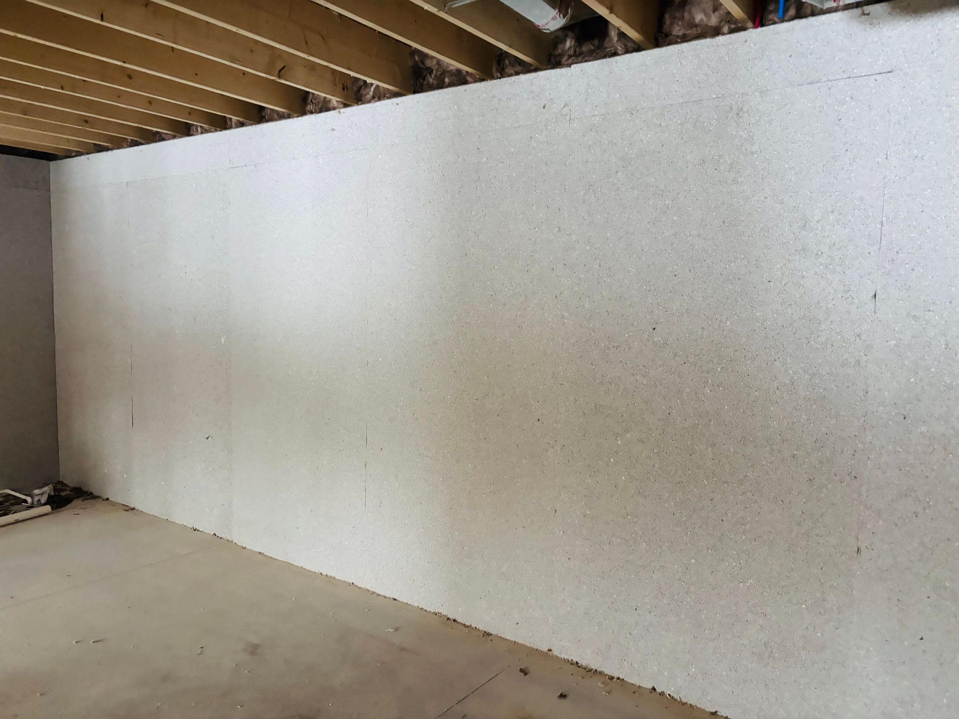 Interior view of a room with unfinished walls. White insulation panels cover the walls, with exposed wood ceiling beams above. Concrete floor.