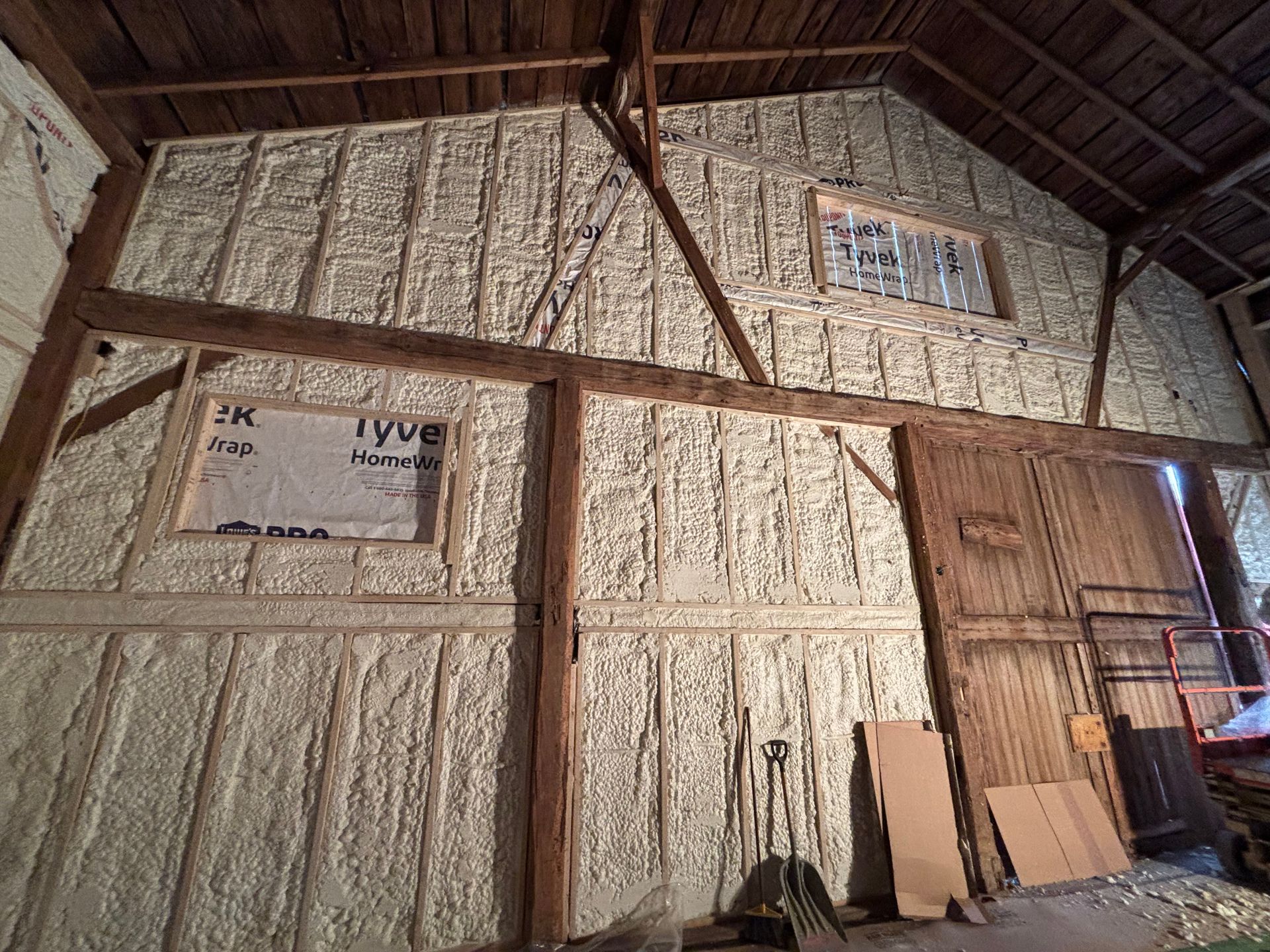 Interior of a barn with walls insulated with light-colored spray foam, wooden beams, and a window.