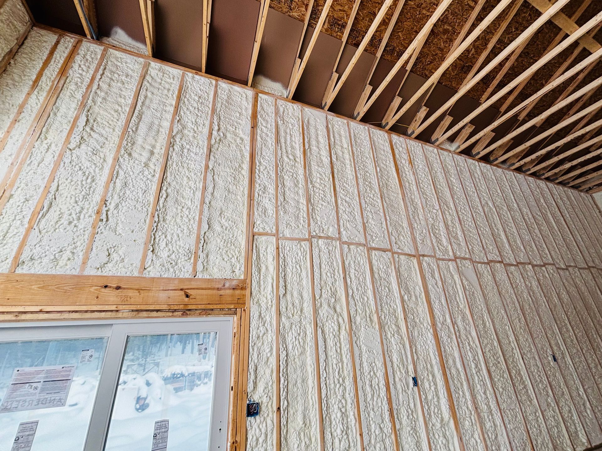 Interior wall with spray foam insulation between wooden studs, partially covering an open ceiling.