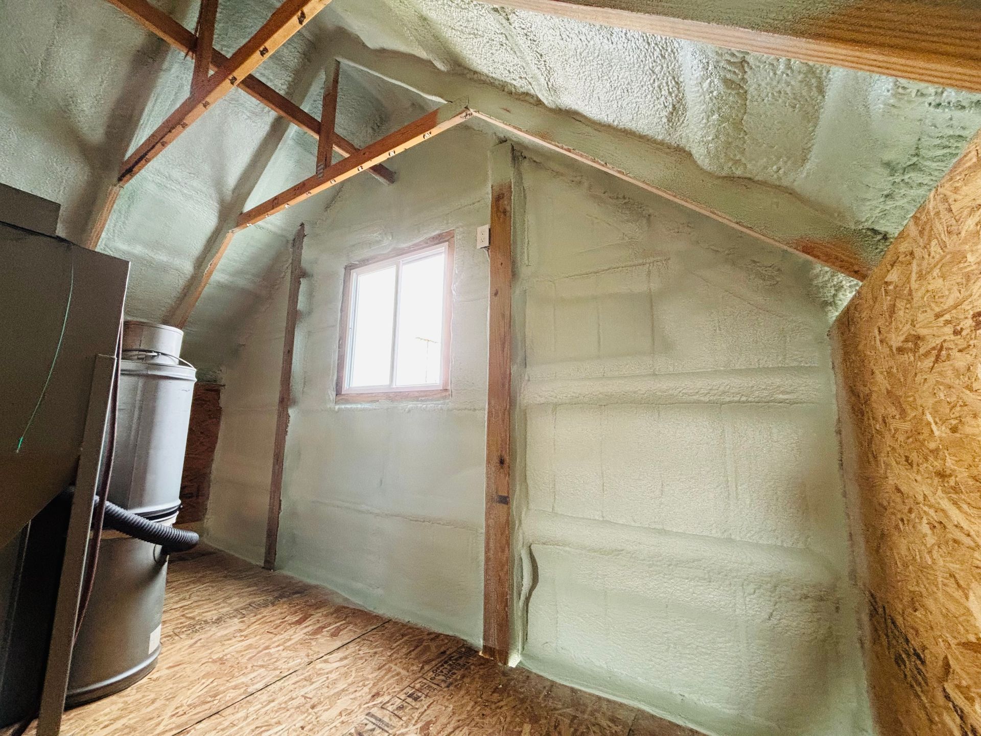 Interior view of a small attic space insulated with light green spray foam, with a window and a water heater visible.
